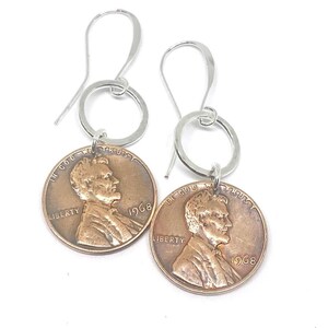 May include: A pair of silver hoop earrings with a 1968 penny charm attached to each hoop. The penny features a profile of Abraham Lincoln and the words "In God We Trust", "Liberty", and "1968".