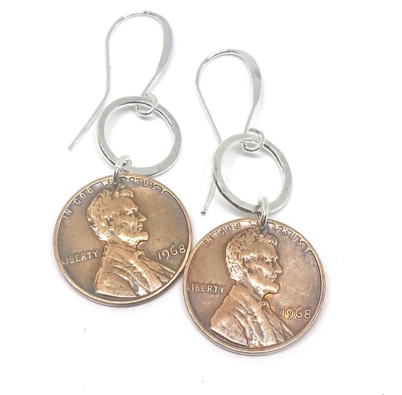 May include: A pair of silver hoop earrings with a 1968 penny charm attached to each hoop. The penny features a profile of Abraham Lincoln and the words "In God We Trust", "Liberty", and "1968".