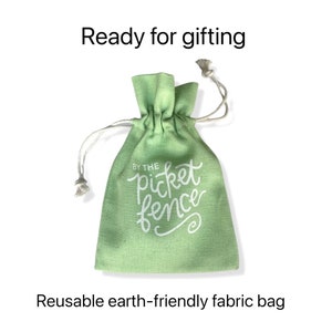 May include: A light green reusable fabric bag with a white drawstring. The bag has the text "By the picket fence" printed on it.