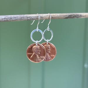 May include: A pair of silver hoop earrings with a 1974 US penny attached to each hoop.