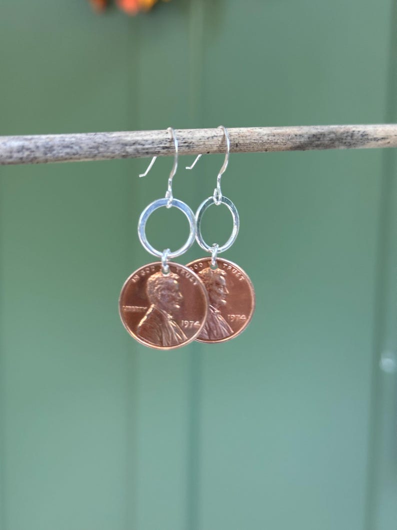 May include: A pair of silver hoop earrings with a 1974 US penny attached to each hoop.