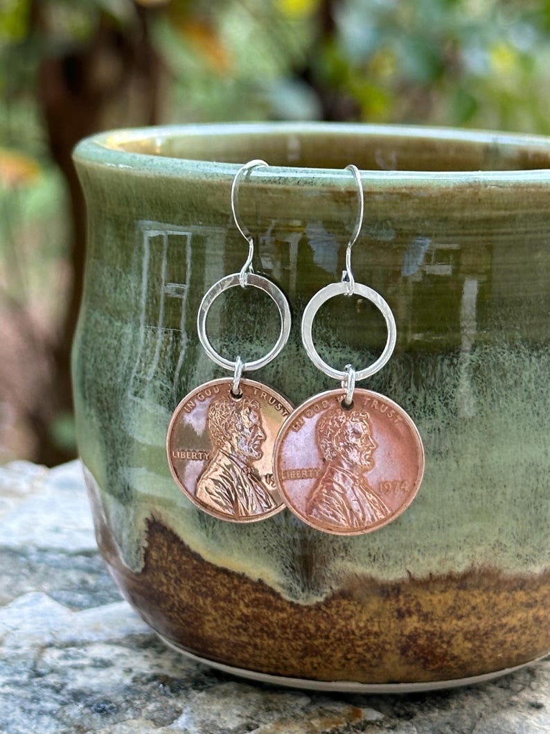 May include: A pair of silver hoop earrings with a copper penny charm. The penny is dated 1976 and features the profile of Abraham Lincoln with the inscription "In God We Trust" and "Liberty".