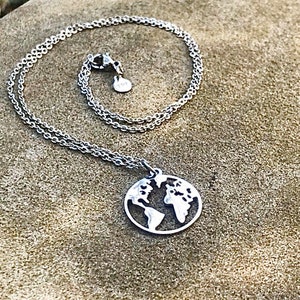 Puede incluir: Un collar de plata con una cadena delicada y un colgante con forma de la Tierra. El colgante está recortado para mostrar los continentes y los océanos.