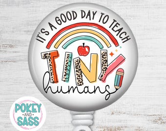 May include: A circular badge reel featuring the text IT'S A GOOD DAY TO TEACH TINY HUMANS. The design includes a colorful rainbow, a red apple, a ruler, and a pencil. The letters are decorated with leopard print and solid orange and yellow colors.