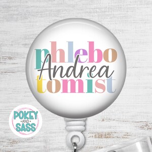 May include: A round white badge reel featuring the colorful text phlebotomist with the name Andrea written in a dark gray script font across the center. The badge reel has a white plastic housing and a retractable cord mechanism for identification.