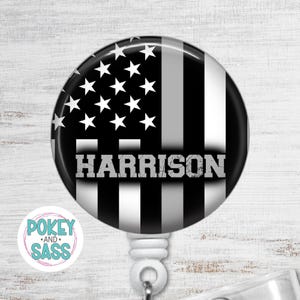 May include: A circular badge reel featuring a black and white American flag design with a field of stars on the left and vertical stripes on the right. The name HARRISON is printed in a bold, distressed gray font across the center of the flag.