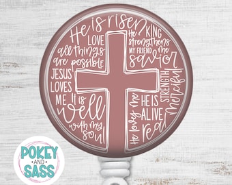 May include: A circular badge reel featuring a dusty rose background with a white outline of a cross. The design includes white handwritten text such as He is risen, King, Love, all things are possible, Jesus loves me, It is well with my soul, and He is alive.