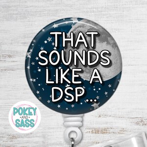 May include: A round, navy blue badge reel with the text "THAT SOUNDS LIKE A DSP..." in white. The design includes a crescent moon and white stars. The badge reel has a white plastic clip and a "Pokey and Sass" logo.
