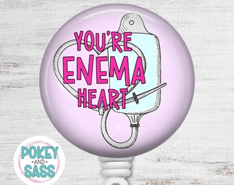 May include: A round badge reel featuring a light purple background with a black and white line drawing of an enema bag and tubing shaped into a heart. The text YOU'RE ENEMA HEART is printed in bold pink letters across the center of the design.