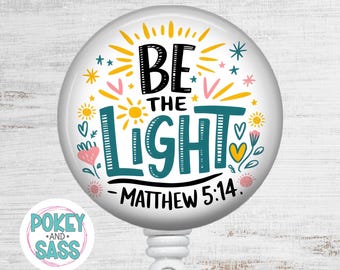 May include: A circular badge reel featuring a white background with the black and teal text BE THE LIGHT and MATTHEW 5:14. The design includes yellow sunbursts, small stars, and pink and yellow hearts and flowers surrounding the central text.