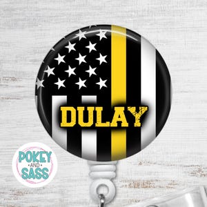 May include: A circular badge reel featuring a stylized American flag design with black and white stripes, a field of white stars on a black background, and a single vertical yellow stripe. The name DULAY is printed in bold yellow block letters.