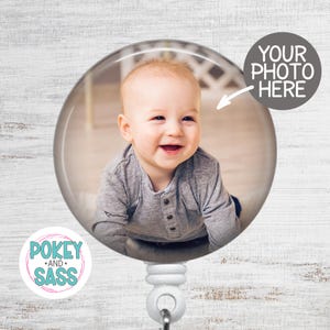 May include: A clear, round badge reel with a smiling baby photo inside. The badge reel has a white base and a retractable cord. Text on the badge reel says "YOUR PHOTO HERE" and "POKEY AND SASS".
