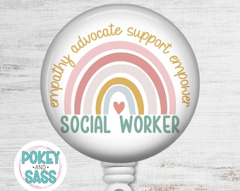 May include: A circular badge reel featuring a white background with a pastel rainbow graphic. The text reads empathy advocate support empower in gold and SOCIAL WORKER in teal. A small red heart sits below the rainbow. A logo reads POKEY AND SASS.