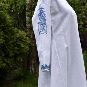 May include: A white linen tunic with blue embroidered Celtic knot designs on the sleeve and cuff. The tunic is long and flowing, with a simple neckline.