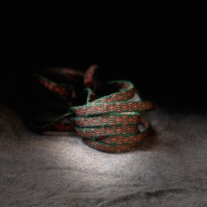 May include: Coiled orange and green patterned cord on a gray fabric surface. The cord appears to be woven with a repeating geometric design. The image is set against a dark background, highlighting the texture and color of the cord.