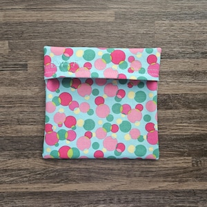 Puede incluir: Una bolsa de aperitivos reutilizable cuadrada con cierre de solapa. La bolsa presenta un fondo azul claro con un patrón de círculos rosas, verdes y amarillos. La bolsa está hecha de tela y está diseñada para guardar aperitivos.