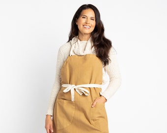May include: A woman wearing a white sweater and a mustard yellow apron with a white tie. The apron has two large pockets.