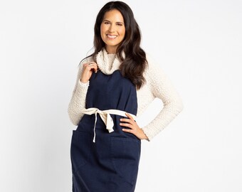 May include: A woman wearing a navy blue apron with a white tie and a white sweater. She is smiling and looking at the camera.