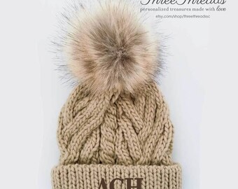 May include: A beige knitted beanie with a brown faux fur pom pom and the letters "ACH" embroidered on the front.