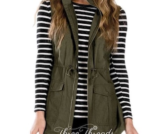 May include: A green utility vest with a zipper closure, two front pockets, and a drawstring at the waist. The vest is worn over a black and white striped long-sleeved shirt.