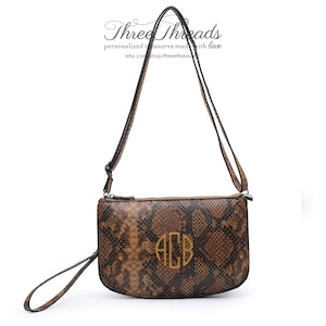 Może przedstawiać: Brązowa torebka na ramię ze wzorem skóry węża i złotym monogramem "ACB" z przodu. Torebka ma zamek błyskawiczny i odpinany pasek.