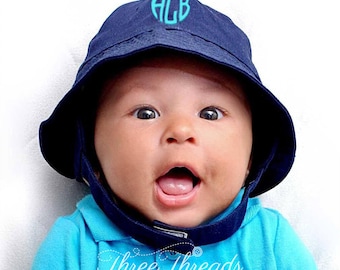 May include: A baby wearing a blue and white sun hat with the monogram "ACB" embroidered on the front. The hat has a chin strap and a brim that covers the baby's ears. The baby is wearing a turquoise polo shirt. The text "Three Threads" is visible at the bottom of the image.