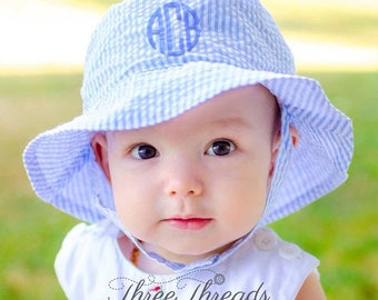 May include: A baby wearing a blue and white striped sun hat with a blue monogram "ACB" on the front. The hat has a wide brim and a chin strap. The baby is looking at the camera.
