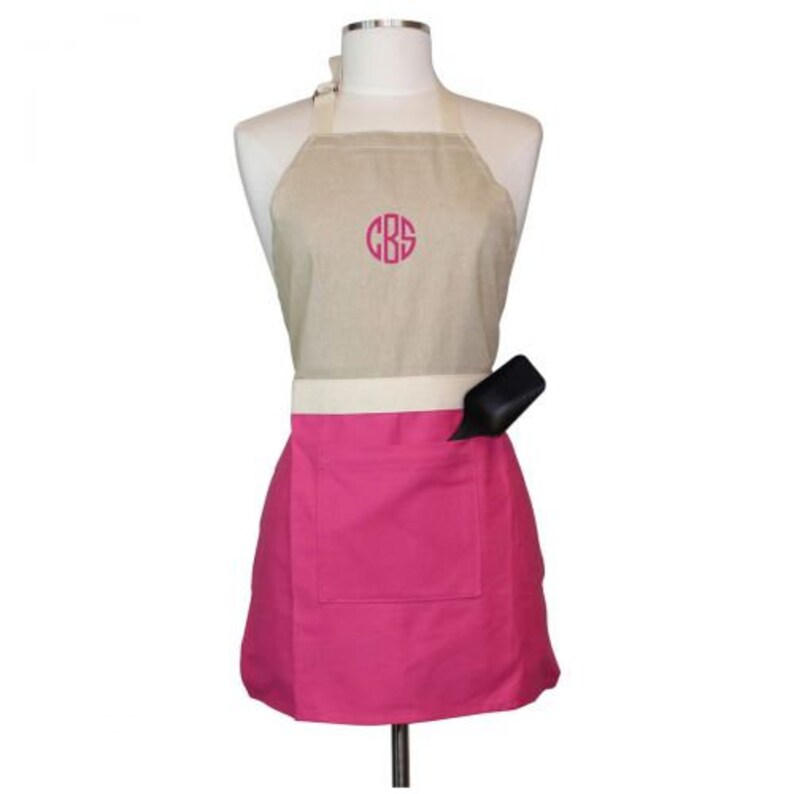 Op de afbeelding: Een beige en roze schort met een grote zak en een gereedschapshouder. Het schort heeft een monogram "CBS" in roze op de bovenste helft.