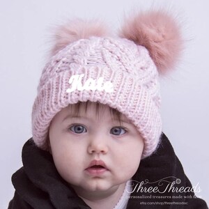 May include: A pink knitted baby hat with two fluffy pom-poms on top. The hat has the name "Kate" embroidered on the front in white.