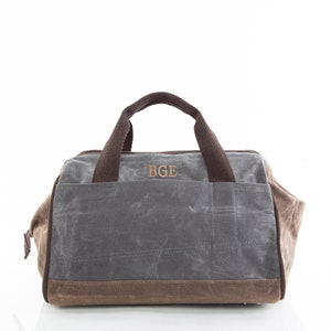 Puede incluir: Una bolsa de lona gris con ribetes de cuero marrón y un asa de cuero marrón. La bolsa tiene cierre de cremallera y un bolsillo grande en la parte delantera. Las iniciales "BGE" están bordadas en la parte delantera de la bolsa.