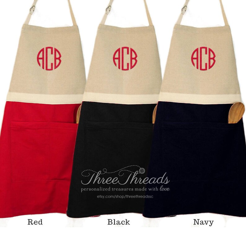 Op de afbeelding: Drie schorten met een witte bovenkant en een zak, en een onderkant in rood, zwart en marineblauw. Elke schort heeft een witte cirkel met de letters "ACB" geborduurd in rood.
