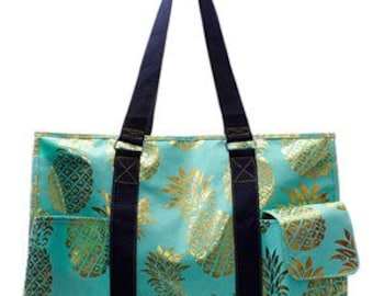 May include: A teal blue tote bag with a gold pineapple print. The bag has two navy blue handles and a smaller pocket on the front.
