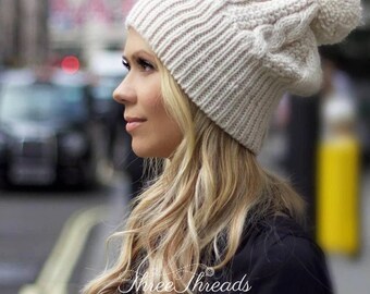 May include: A white knitted beanie with a pom pom on top. The beanie has a cable knit pattern and is worn by a person with long blonde hair.