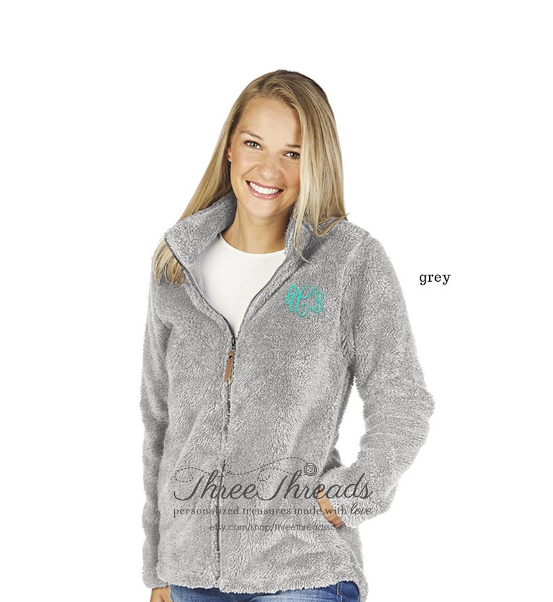 May include: A grey fleece jacket with a zip fastening and a teal monogram on the chest. The jacket has a soft, fuzzy texture. The text "Three Threads" is visible on the bottom of the jacket.