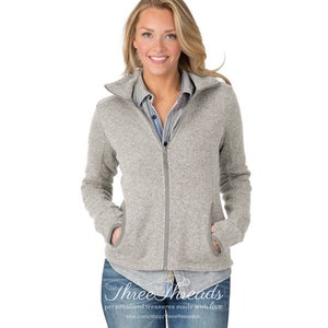 May include: A gray zip-up fleece jacket with a stand-up collar and thumbholes. The jacket has a zipper closure and two pockets. The model is wearing the jacket with a blue button-down shirt and blue jeans.