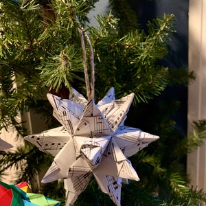 Pode incluir: Um enfeite de estrela de origami branco pendurado em uma árvore de Natal artificial verde. A estrela é feita de papel com um padrão de notas musicais em preto e branco.