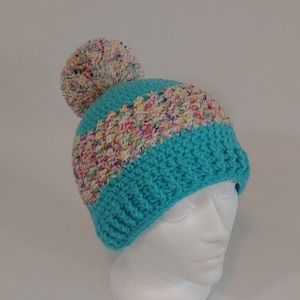 May include: A handmade turquoise crochet beanie with a multicolored yarn band and a large pom-pom on top. The hat is displayed on a white mannequin head, showcasing its texture and design. This winter hat is perfect for cold weather.