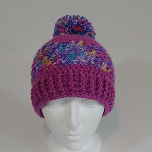 May include: A handmade, magenta crochet beanie with a ribbed brim and a multi-colored pom-pom. The top section features a colorful, textured pattern with blue, yellow, and purple hues. The hat is displayed on a white mannequin head.