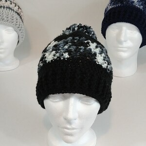 May include: Three handmade crochet beanies on white mannequin heads. The front beanie is black with white and gray accents and a black pom-pom. The other beanies are gray and navy blue with white accents. The beanies are displayed on a white background.