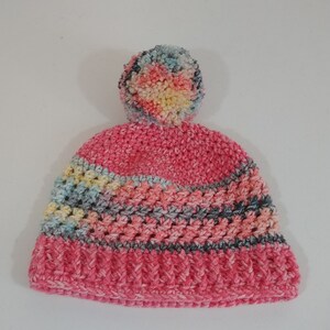 May include: A handmade pink crochet hat with a multi-colored pom-pom on top. The hat features a ribbed brim and a striped pattern in shades of pink, blue, yellow, and gray. This cozy winter hat is perfect for staying warm.