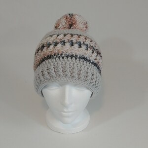 May include: A handmade crochet beanie in shades of gray, beige, and black, topped with a fluffy pom-pom. The hat features a ribbed brim and a textured stitch pattern on the crown, perfect for cold weather.