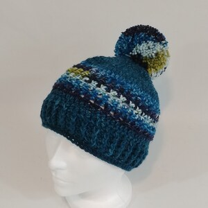 May include: A hand-knitted teal beanie with a large pom-pom. The hat features a ribbed brim and a patterned design in shades of blue, white, and green. The pom-pom is approximately 4 inches in diameter.