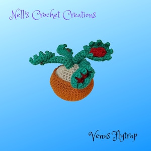 May include: A crocheted Venus flytrap plant with green leaves and red inner parts. The plant is in a brown pot with a white base. The text "Nell's Crochet Creations" is at the top of the image and "Venus Flytrap" is at the bottom.
