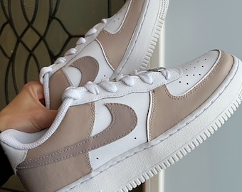 May include: White and beige custom-painted Nike Air Force 1 sneakers with a contrasting swoosh logo. The shoes have a white sole with a textured tread.