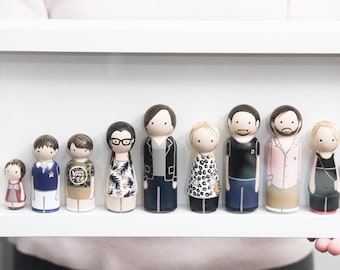 May include: A white frame with a glass front displays a family of hand-painted wooden peg dolls. The dolls are dressed in various outfits, including a floral dress, a black leather jacket, and a blue and white striped shirt. There is also a small dog doll.