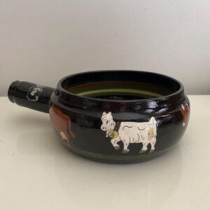 May include: A black ceramic pot with a single handle, featuring hand-painted illustrations of a white goat with a bell and a brown cow. The exterior has horizontal green and red stripes near the rim and base, with a glossy finish throughout.
