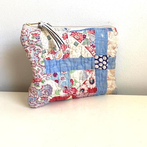 May include: A patchwork fabric pouch with a gold zipper and a striped fabric pull. The pouch features a variety of floral and geometric patterns in red, blue, and cream colors. The design is reminiscent of a vintage quilt.