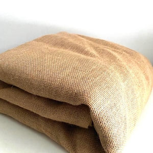 May include: Folded tan burlap fabric. The coarse weave of the fabric is visible, with a natural, earthy tone. The fabric is folded into a neat stack, suggesting it is ready for use in crafts or decor.