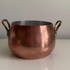 May include: A round copper pot with a polished metallic exterior and a silver-toned interior lining. The vessel features two curved brass handles attached to opposite sides with visible rivets, providing a sturdy grip for this kitchen cookware item.