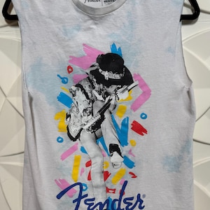 May include: Sleeveless white t-shirt featuring a graphic of a musician playing a guitar, with colorful brushstroke accents. The Fender logo is displayed in blue script. The shirt has a crew neck and the Authentic Hendrix logo.